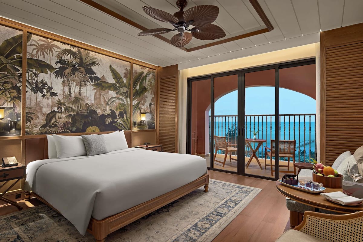 Habitación de hotel con cama, mesita de noche, escritorio con lámpara, silla, mesa de comedor con fruta y un vaso, y balcón con vista al mar.