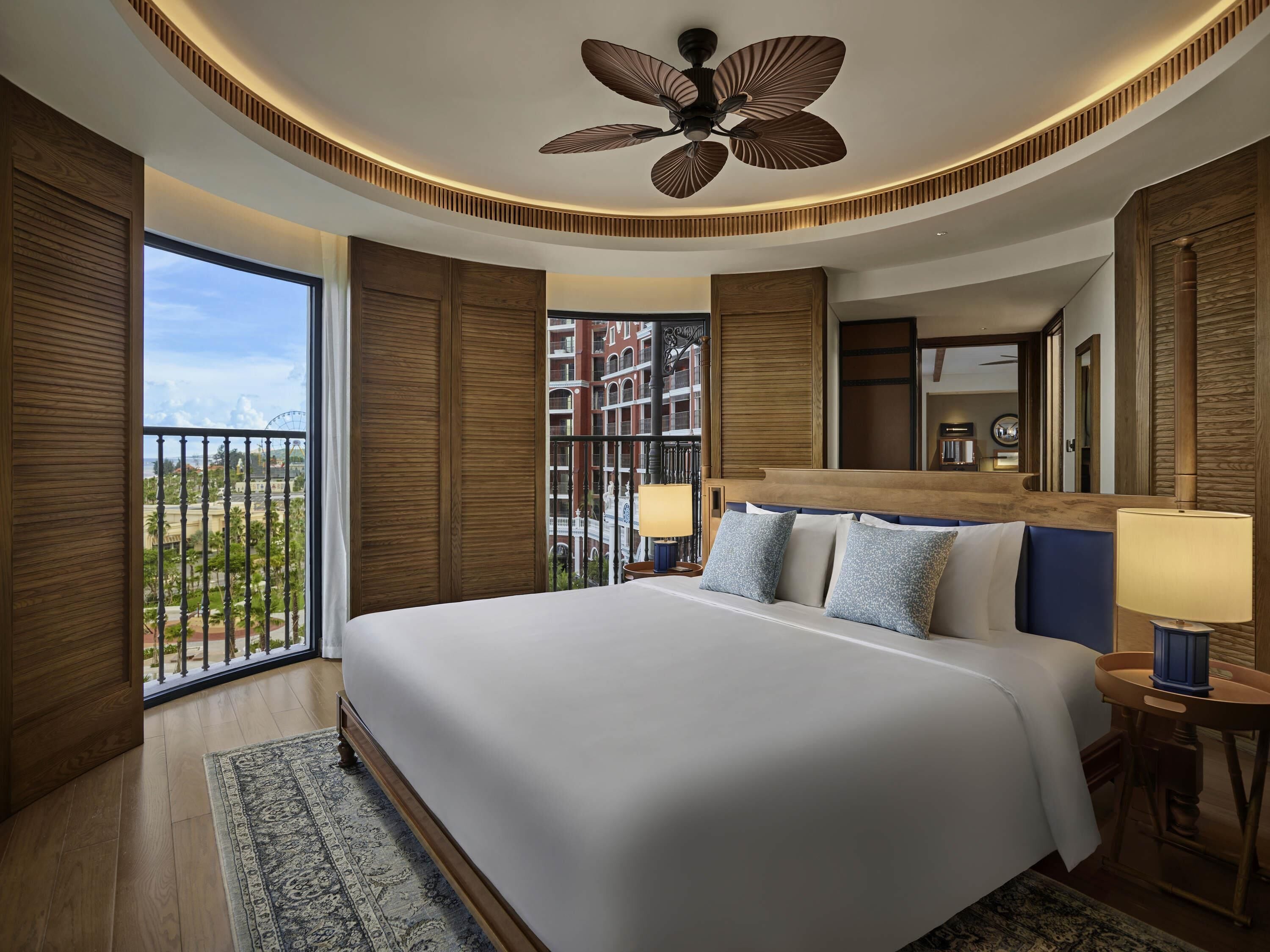 Una habitación de hotel moderna con una cama grande, paneles de madera, un ventilador de techo y un balcón con vistas a edificios y una noria.