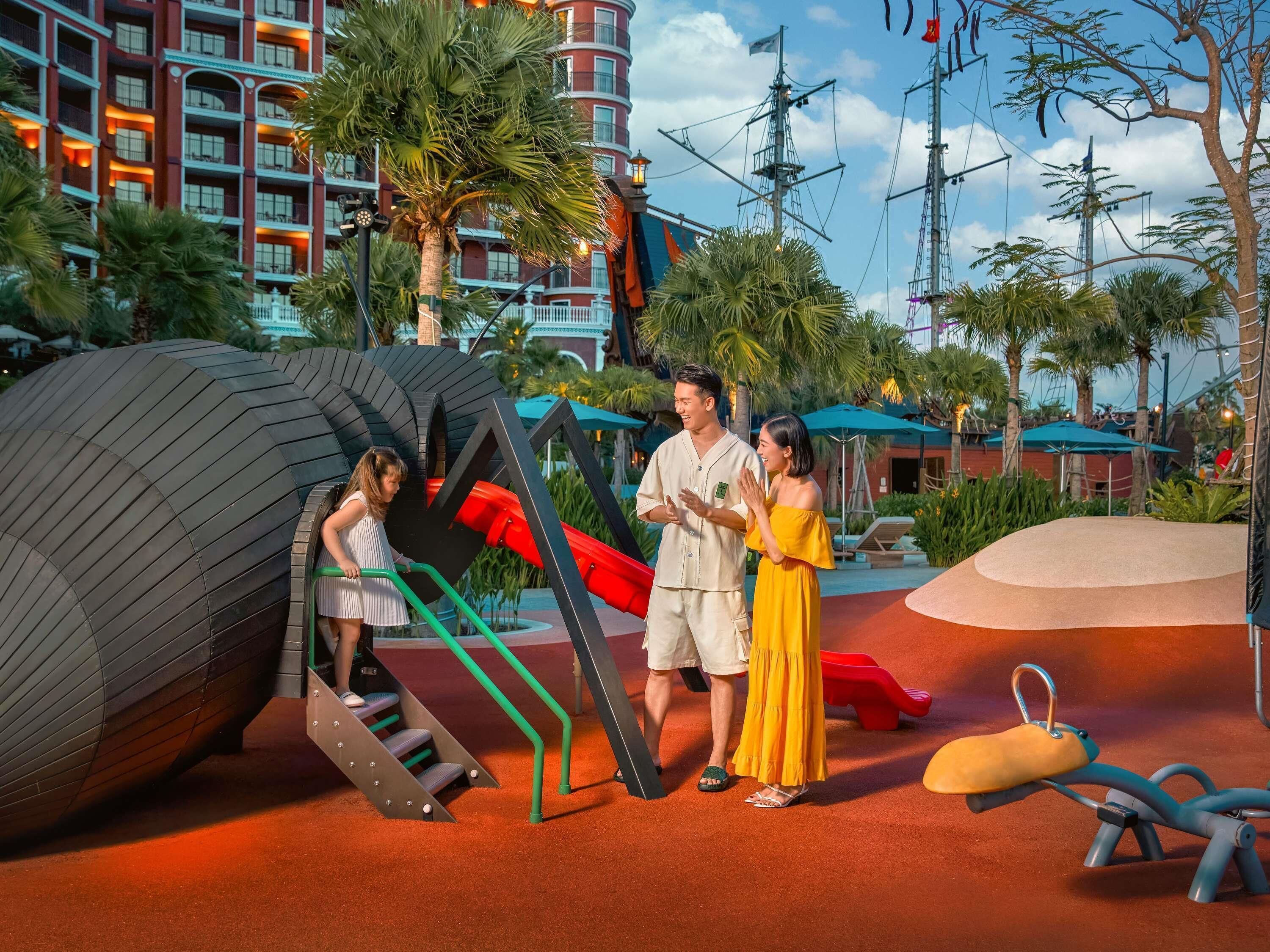A playground with slides and climbing structures, surrounded by palm trees and a building with balconies.