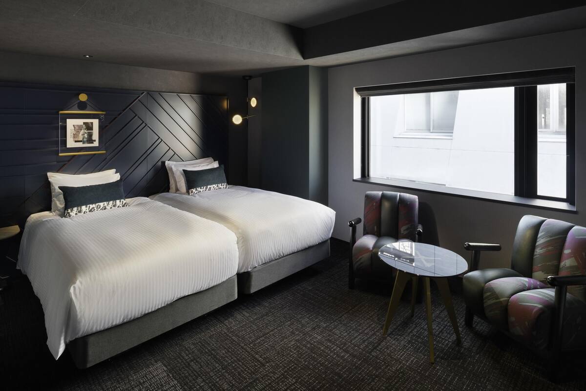 A modern hotel room with a large bed, a seating area with chairs, a small table, and a large window providing a view.