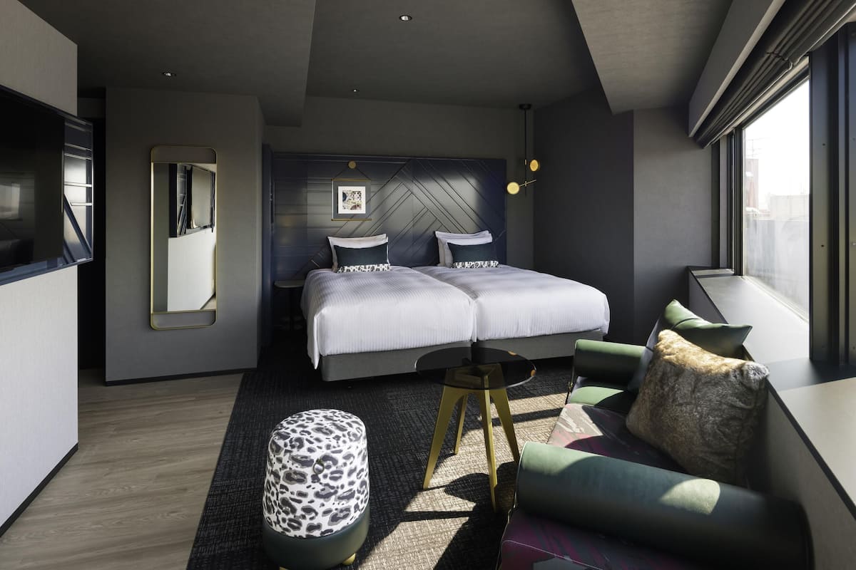 A modern hotel room with a large bed, a sofa, a round coffee table, and a small ottoman.