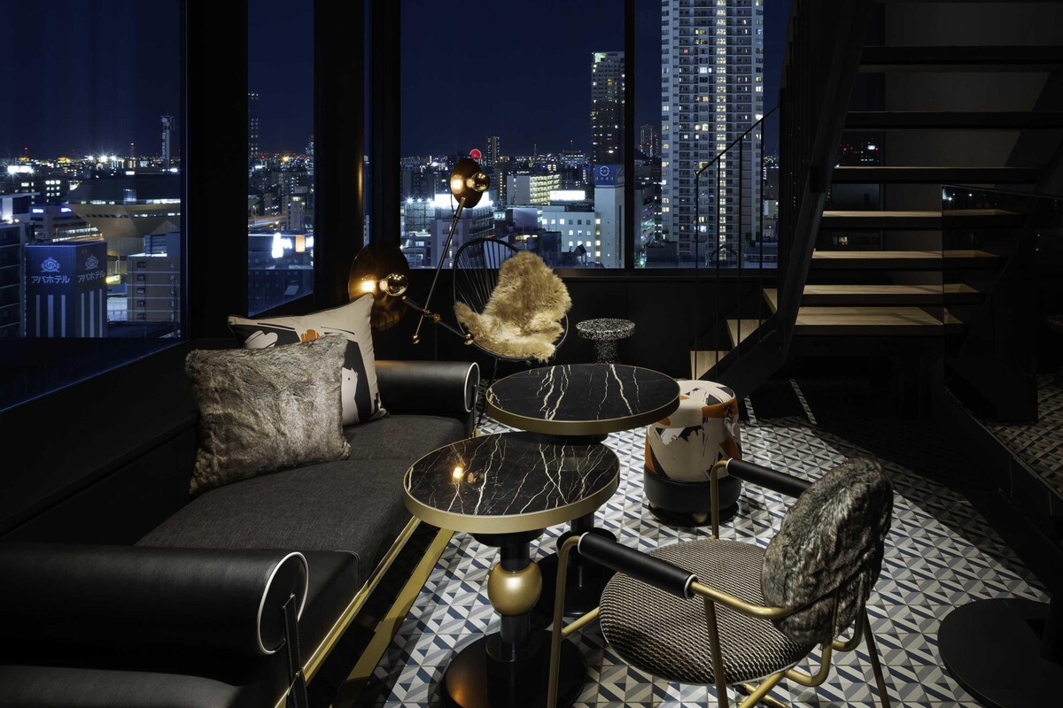 A modern lounge with a sofa, a round table, and a chair, all with gold accents, set against a cityscape view.