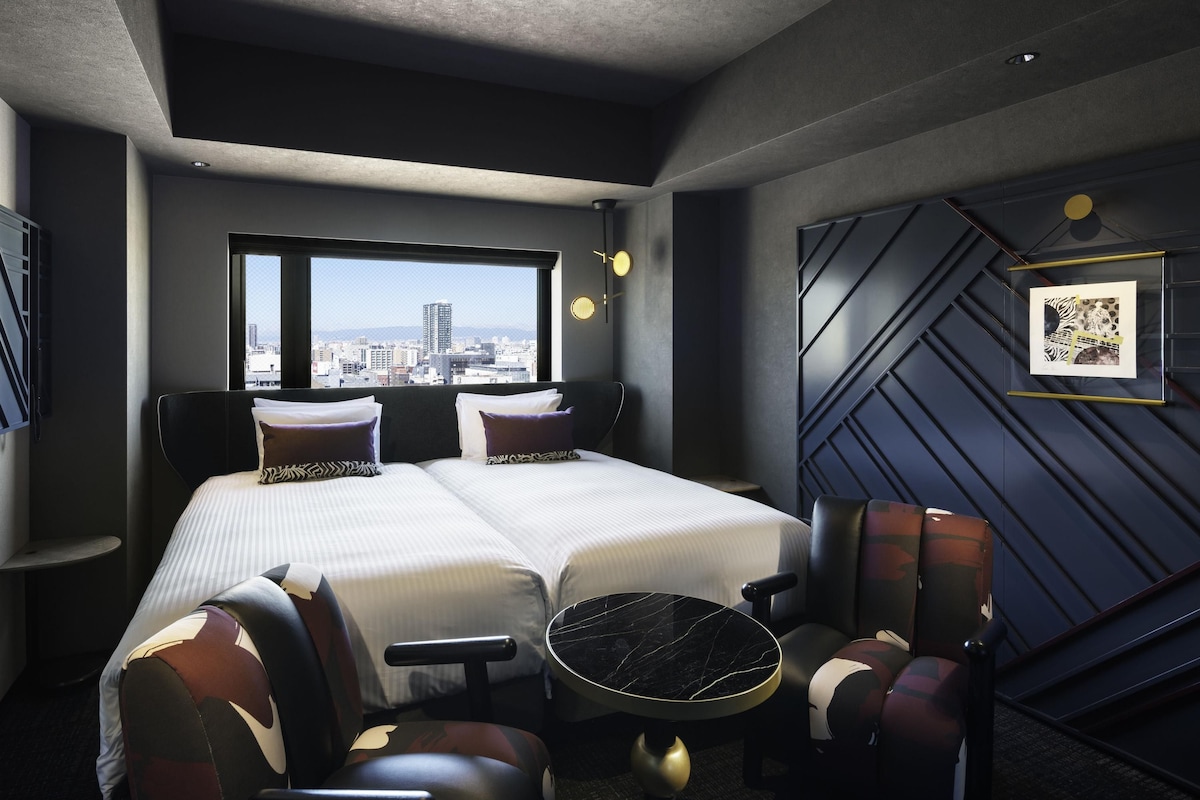 A hotel room with a bed, two chairs, a small table, a window with a city view, and a wall-mounted picture.