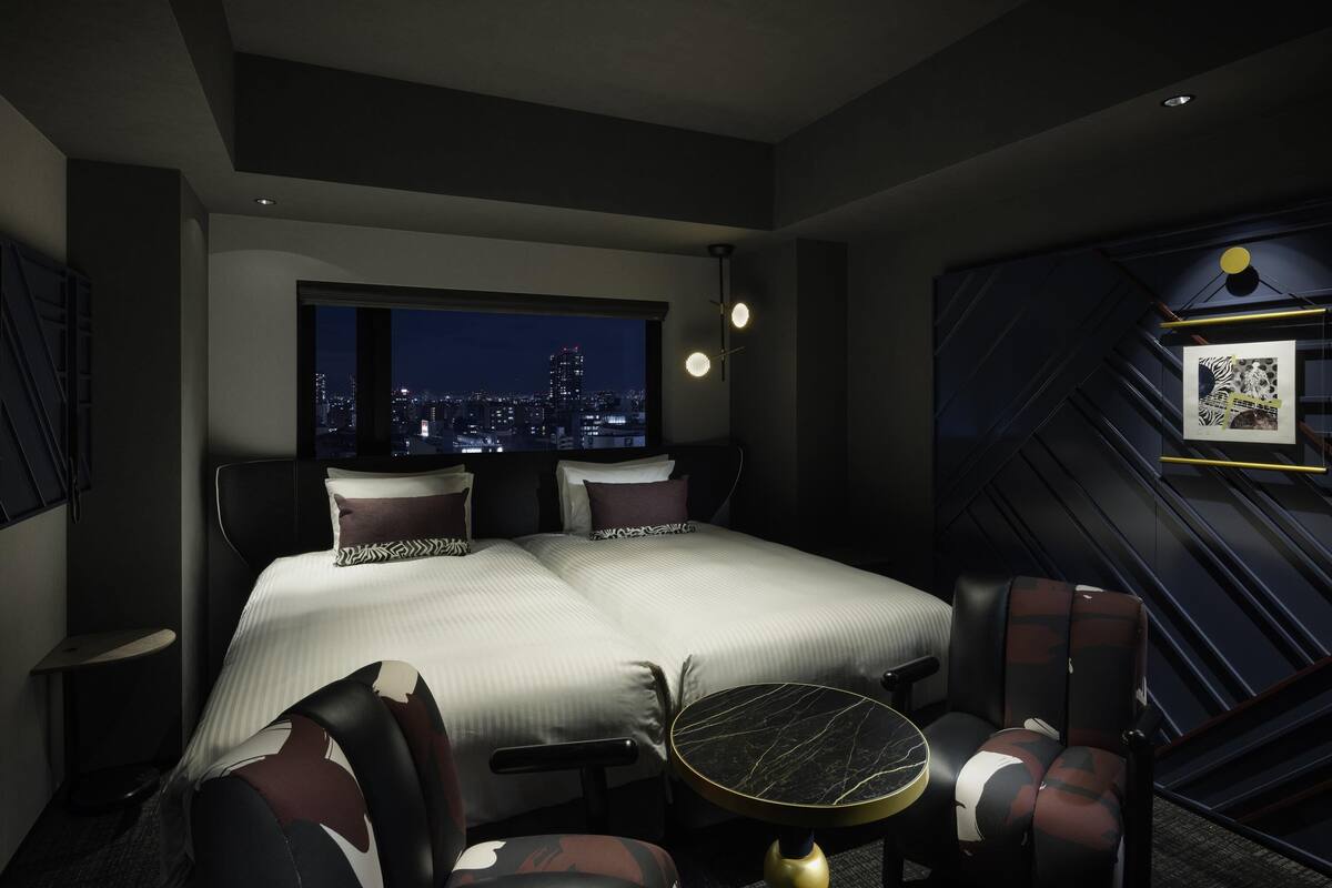 A hotel room with a bed, two chairs, a small table, a city view, and a framed picture on the wall.