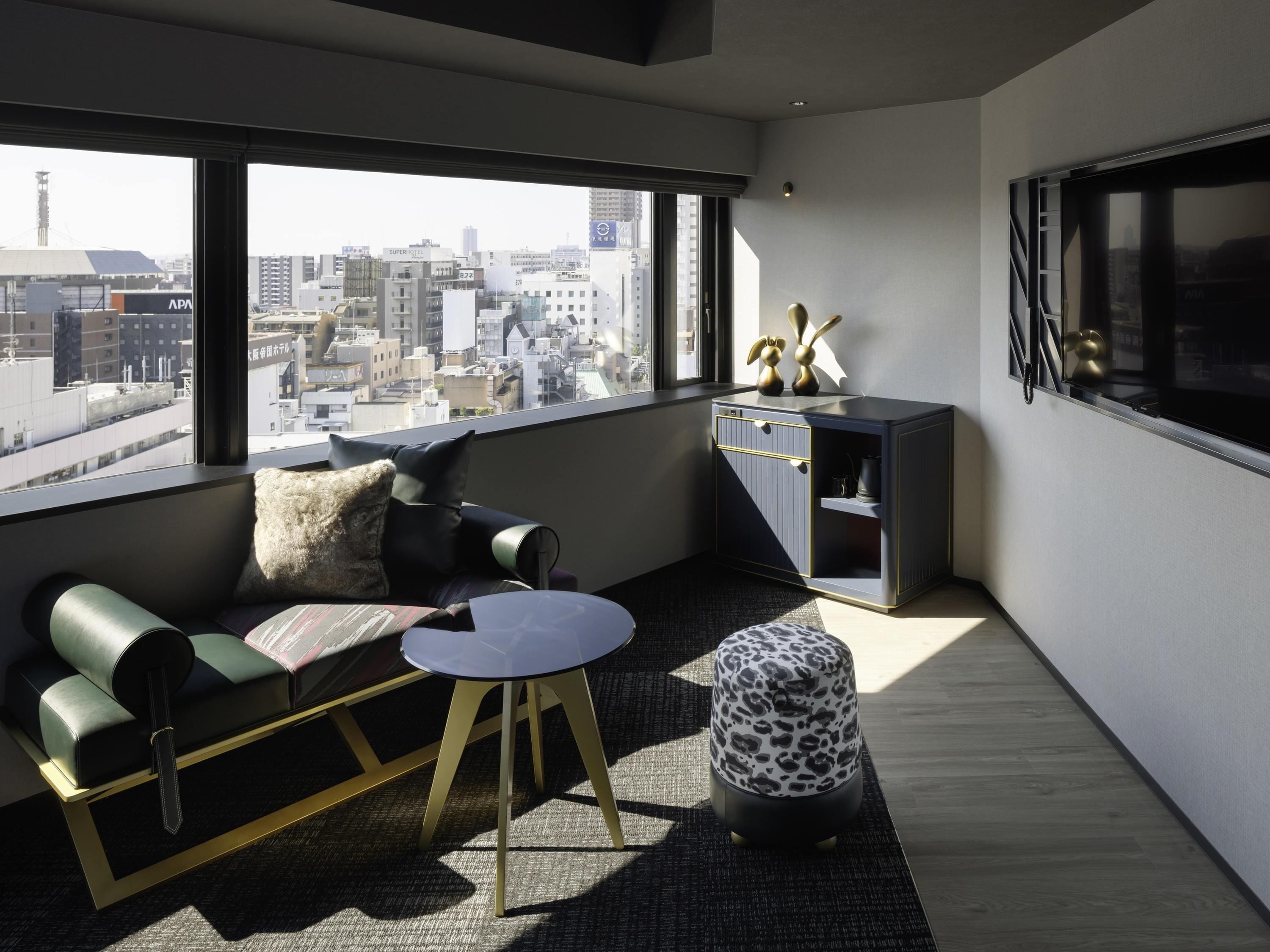 A modern living room with a sofa, ottoman, and coffee table, a cabinet with decorative items, and a large window with a city view.