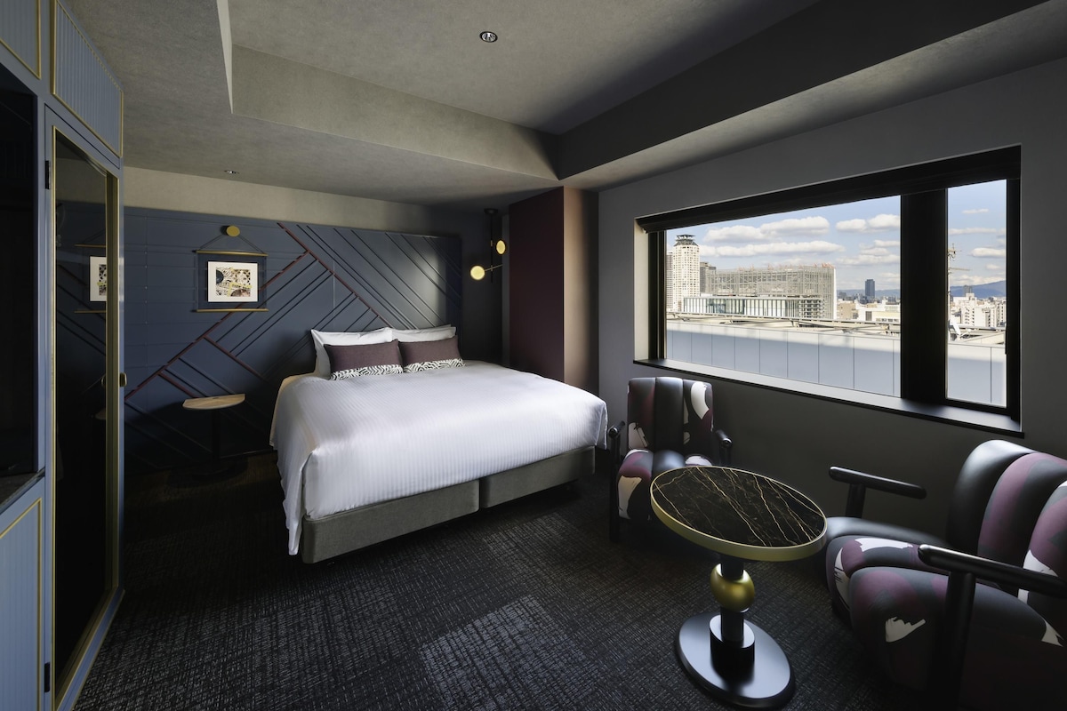 A modern hotel room with a bed, a seating area with a round table, and a large window offering a city view.