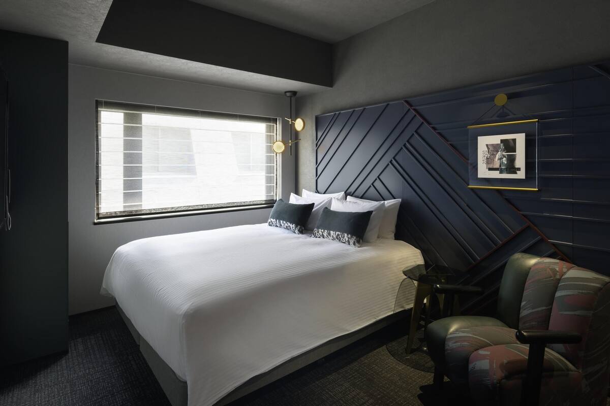 A modern hotel room with a large bed, a desk, and a chair. There is a window with blinds, a wall-mounted picture frame, and a ceiling light fixture.