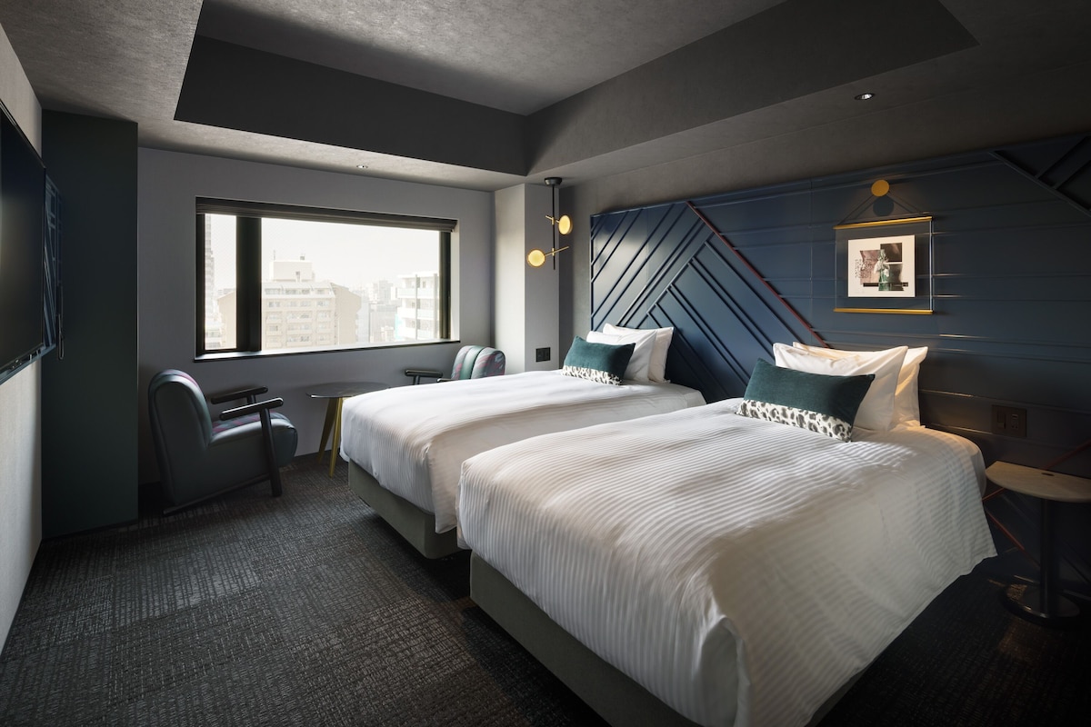 A modern hotel room with two beds, a desk, and a large window offering a city view.