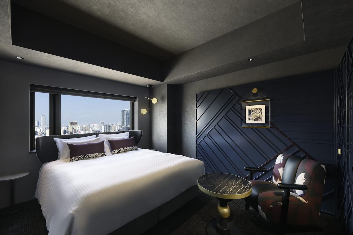 A modern hotel room with a large bed, a desk, a chair, a city view, and a geometric wall design.