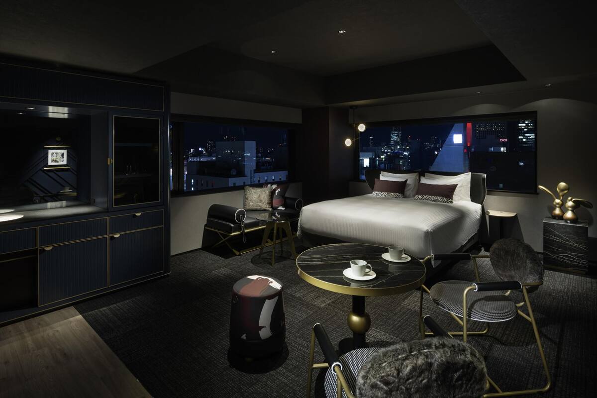 A modern hotel room with a large bed, a sitting area, a small table, and a view of the city at night.
