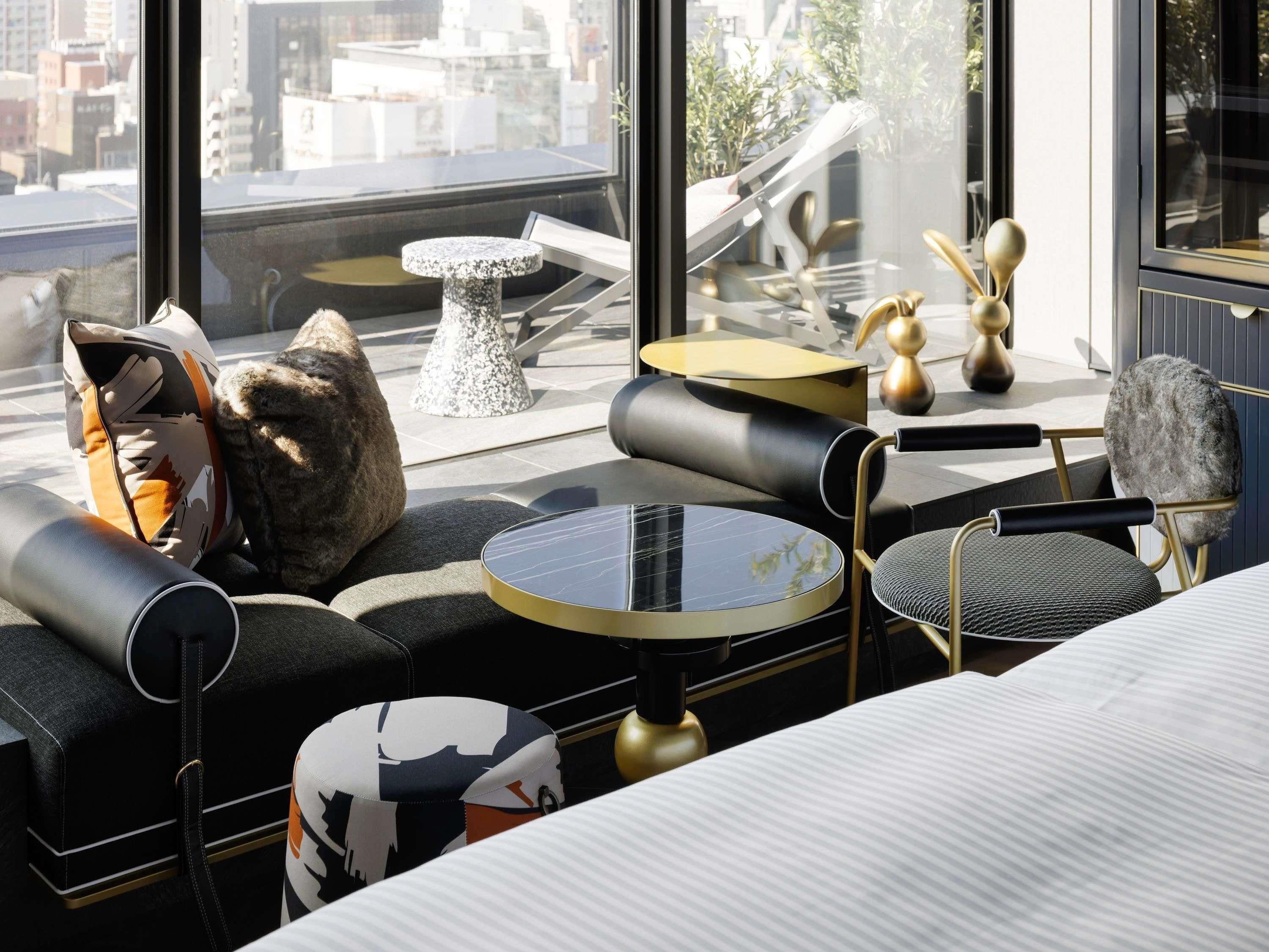 A modern hotel room with a large window, a round table, a sofa, and a chair.