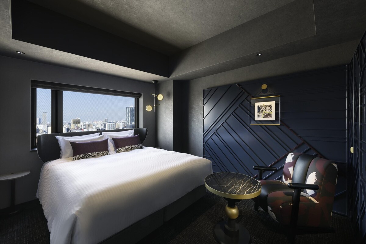 A modern hotel room with a large bed, a desk, a chair, a city view, and a geometric wall design.