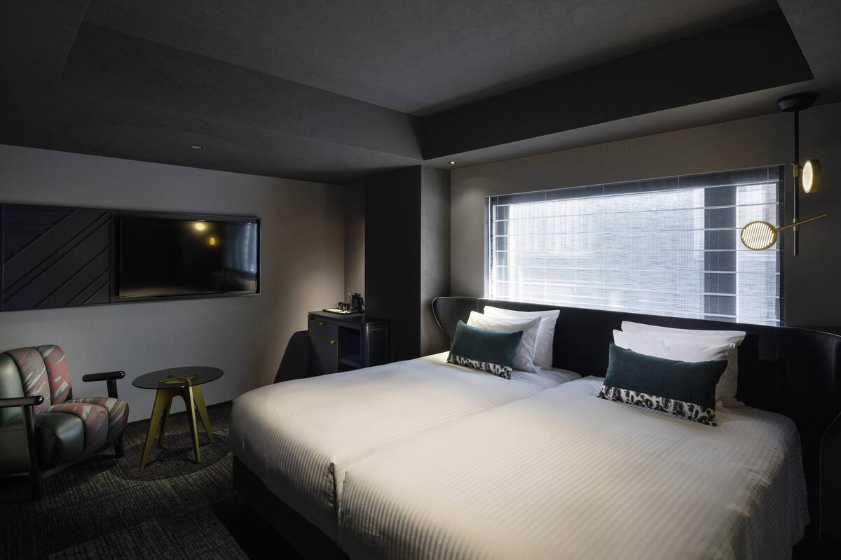 A modern hotel room with a large bed, a desk, a chair, a TV, and a window with blinds.