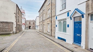 A narrow cobblestone street lined with historic stone buildings, including a church with a dome and a white building with a blue door. - Host & Stay | Apartment 2, High Street (Caernarfon)