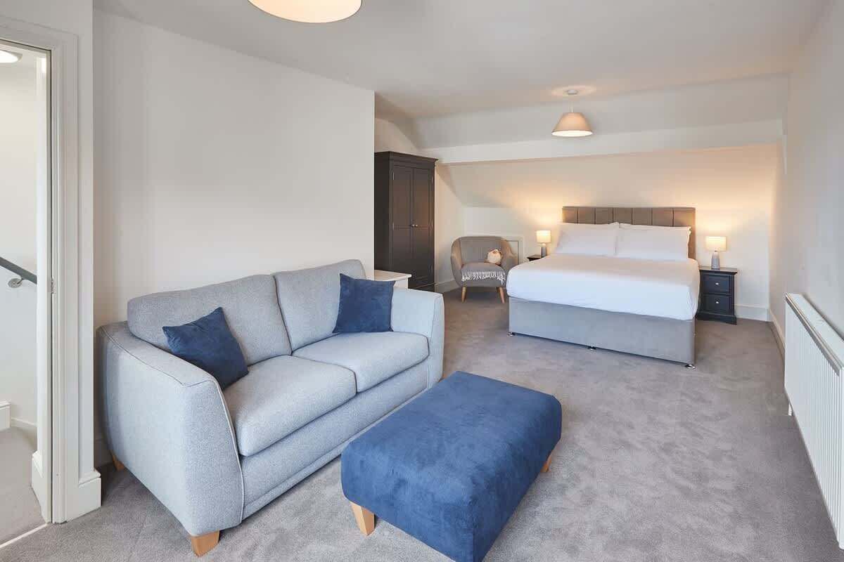 A bedroom with a grey sofa, a blue ottoman, a bed with white bedding, and a chair with a lamp.