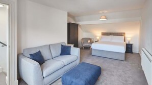 A bedroom with a grey sofa, a blue ottoman, a bed with white bedding, and a chair with a lamp. - Host & Stay | Apartment 2, High Street (Caernarfon)