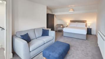 A bedroom with a grey sofa, a blue ottoman, a bed with white bedding, and a chair with a lamp.