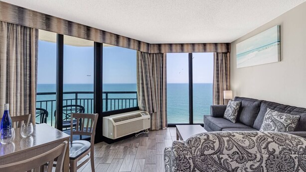 A hotel room with a balcony, a dining table with chairs, a sofa, and a large window offering a view of the sea. - Best Layout/Relaxing OceanfrontSuite (Myrtle Beach)