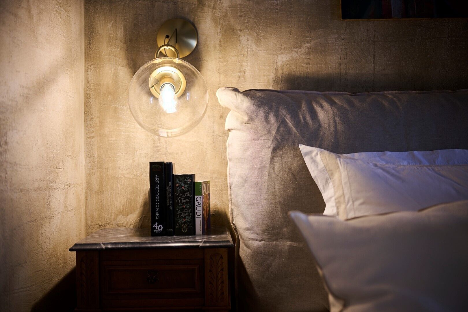 A neatly made bed with white linens, a bedside table with a lamp, and a wall-mounted light fixture.