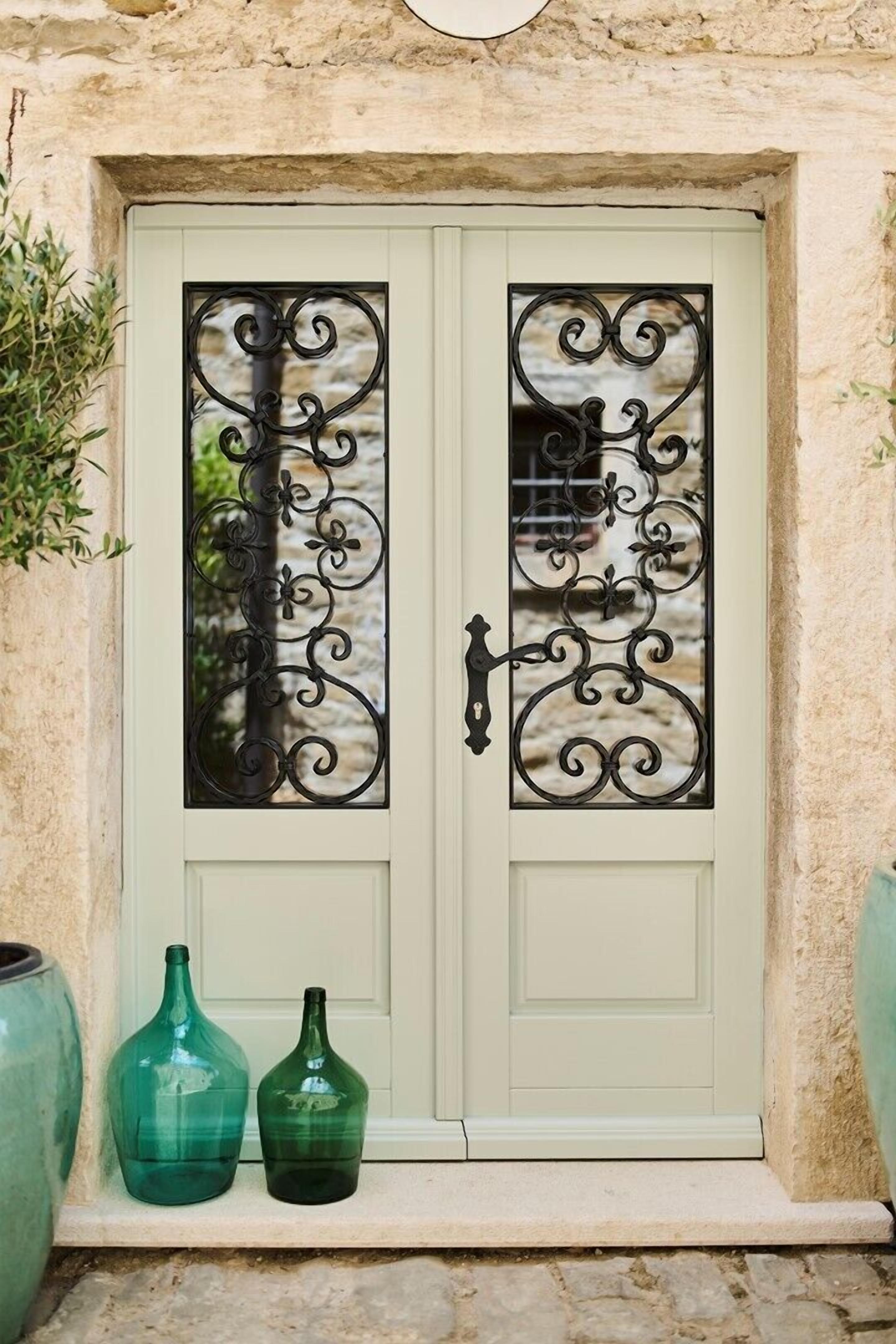 Une porte vert clair ornée de motifs en métal noir et deux bouteilles en verre vert posées sur un rebord en pierre.