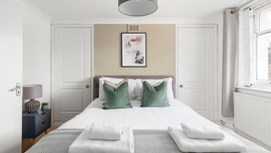 Un dormitorio ordenado con cama, almohadas, una mesita de noche con lámpara y una obra de arte enmarcada en la pared.