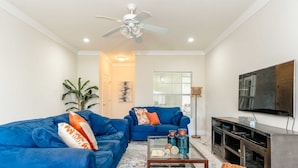 A living room with two blue sofas, a glass coffee table, a wicker chair, a flat-screen TV, and a ceiling fan.
