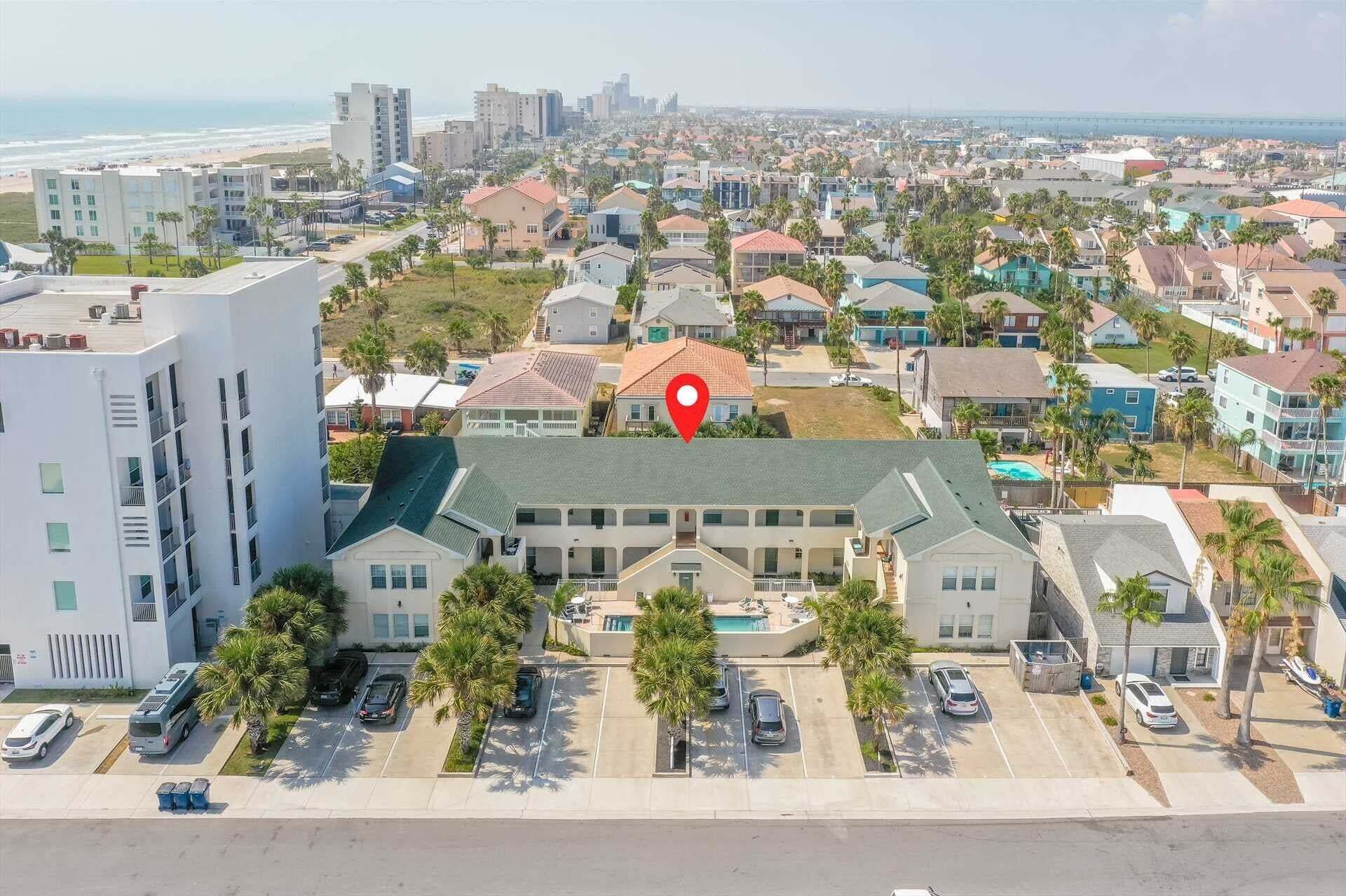 A coastal residential area with a prominent building featuring a large swimming pool, surrounded by parked cars and palm trees.
