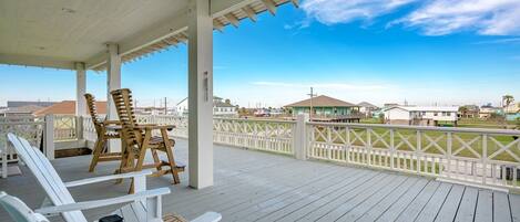 A covered outdoor deck with white chairs and a lantern, overlooking a coastal area with houses and a clear sky.