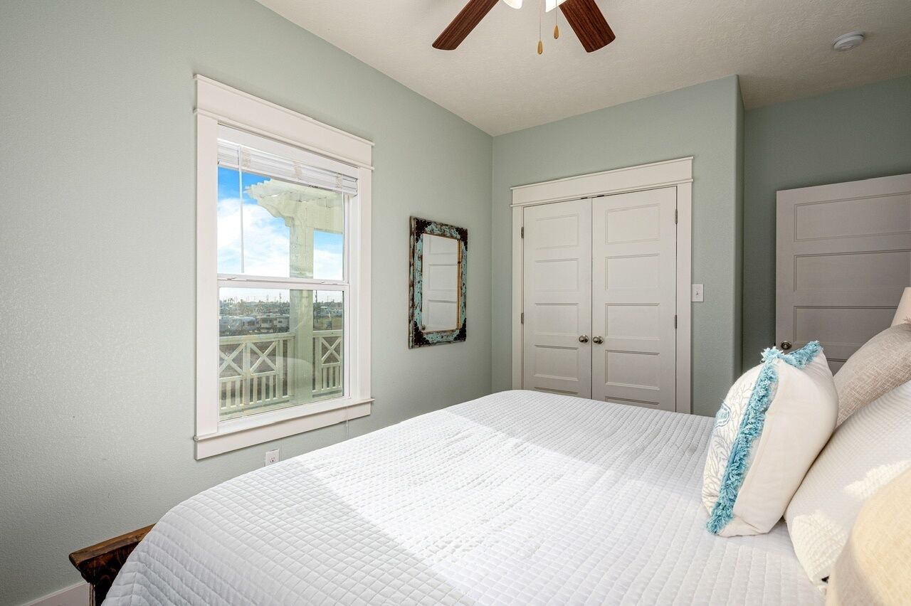 A bedroom with a large bed, a window with a view, a ceiling fan, and a door.