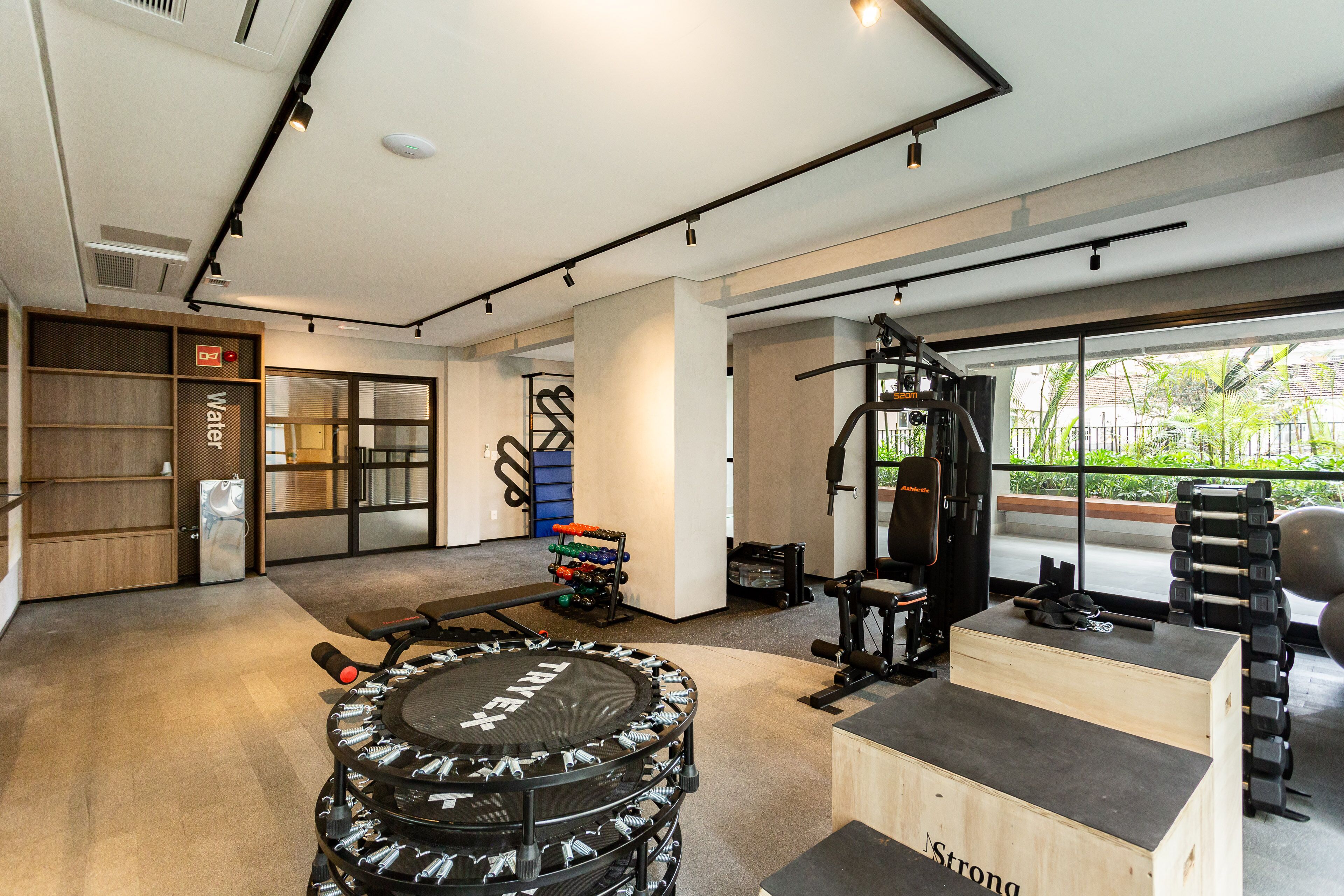 Une salle de sport moderne équipée de divers appareils d’exercice, d’un banc en bois et d’une grande fenêtre donnant sur un paysage verdoyant.