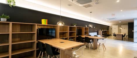 Una biblioteca moderna o sala de lectura con estanterías de madera, sillas negras y traslúcidas, y una larga mesa de madera.