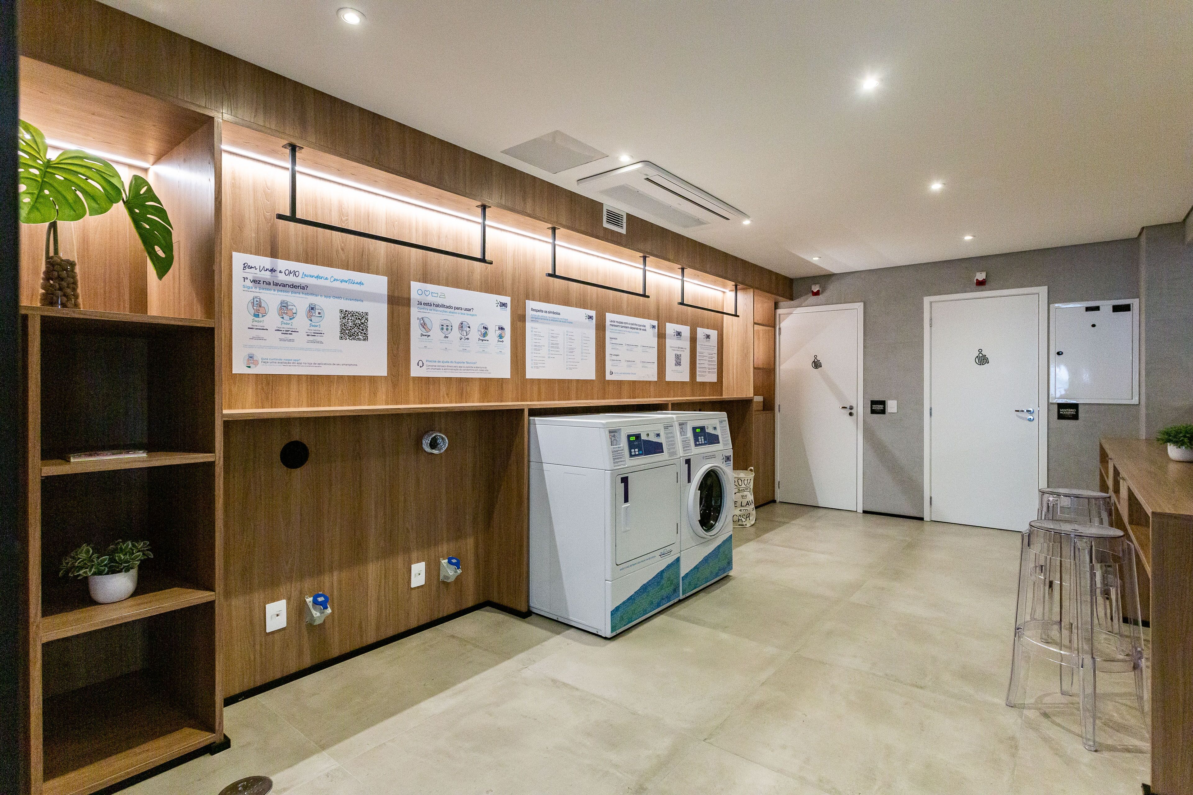 A modern laundry area with a washing machine, clear plastic stools, wooden shelves with plants, and informational posters on the wall.