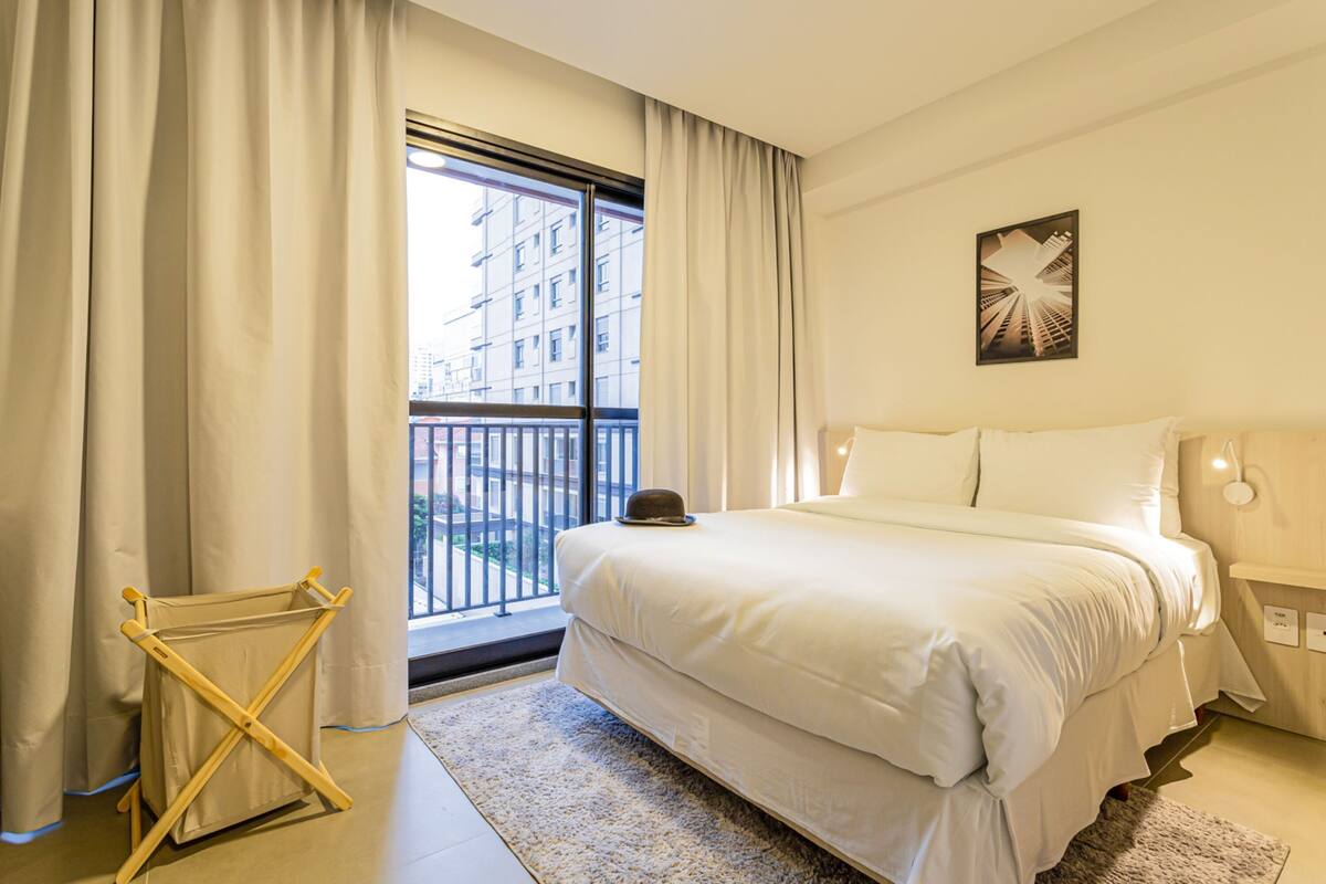 Ein Hotelzimmer mit einem großen Bett, einem Vorhangfenster, einem Hut auf dem Bett und einer Wandleuchte.