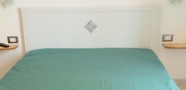 A bed with a teal bedspread, a white headboard, and a decorative diamond-shaped item on the headboard. There are two bedside tables with a remote control on one and a power outlet on the other.