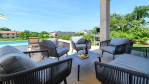 A covered outdoor seating area with wicker furniture and a pool in the background. - Villa Rosmarino Muntrilj with private pool (Muntrilj)