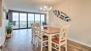 A dining area with a large table and chairs, a chandelier, and a view through large windows. - Fully Renovated Ocean Front Condo at Sea Colony (Sussex County)