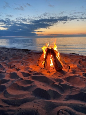 Ein Lagerfeuer an einem sandigen Strand mit Blick auf den Ozean und einem Sonnenuntergang im Hintergrund.