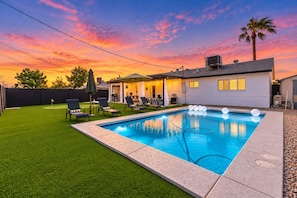 A swimming pool with a patio area, a covered seating area, and a house with a white exterior.
