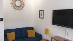 A living room with a blue sofa, a round mirror, a flat-screen TV, a small wooden side table, and a patterned rug.
