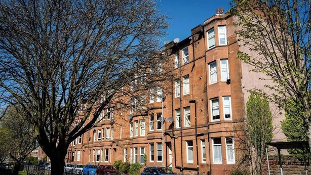 A row of red brick townhouses with multiple windows, a tree in the foreground, and parked cars.