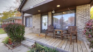Una terraza de madera con dos sillas de madera y una mesa pequeña, una casa de ladrillo con el número 5717, y un pequeño jardín.