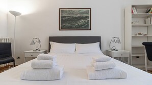Un lit bien fait, avec du linge de lit et des serviettes blanches, une tête de lit grise et un tableau encadré représentant une mer déchaînée accroché au mur.