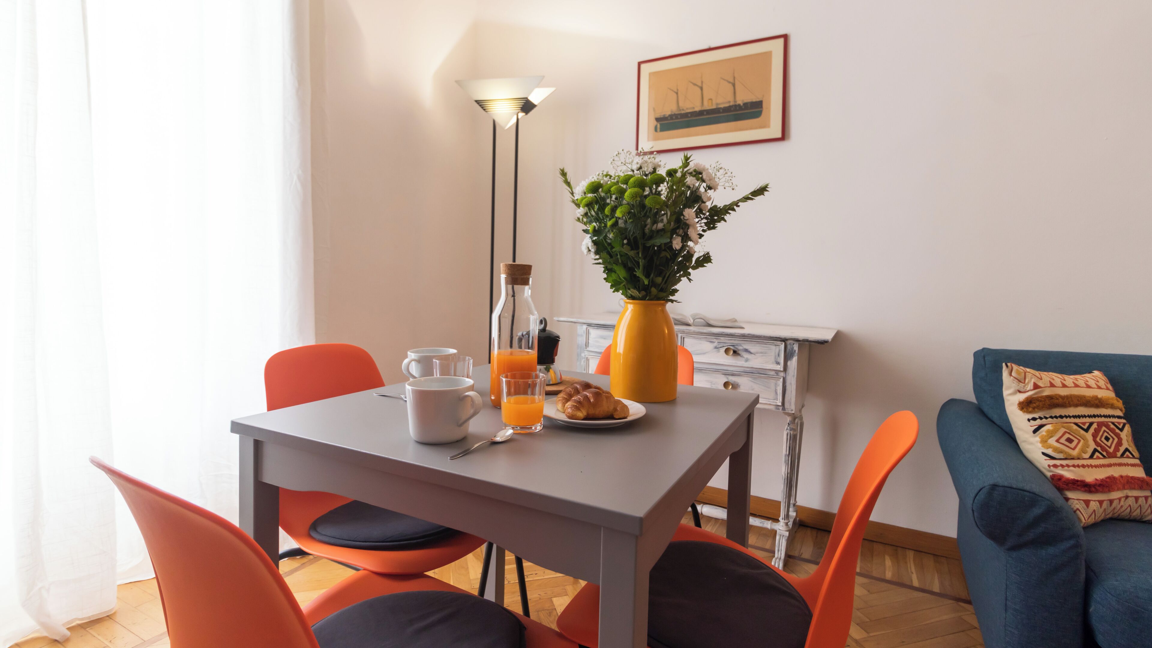 A dining area with a table set for breakfast, including a bottle of orange juice, a croissant, and a vase of flowers.