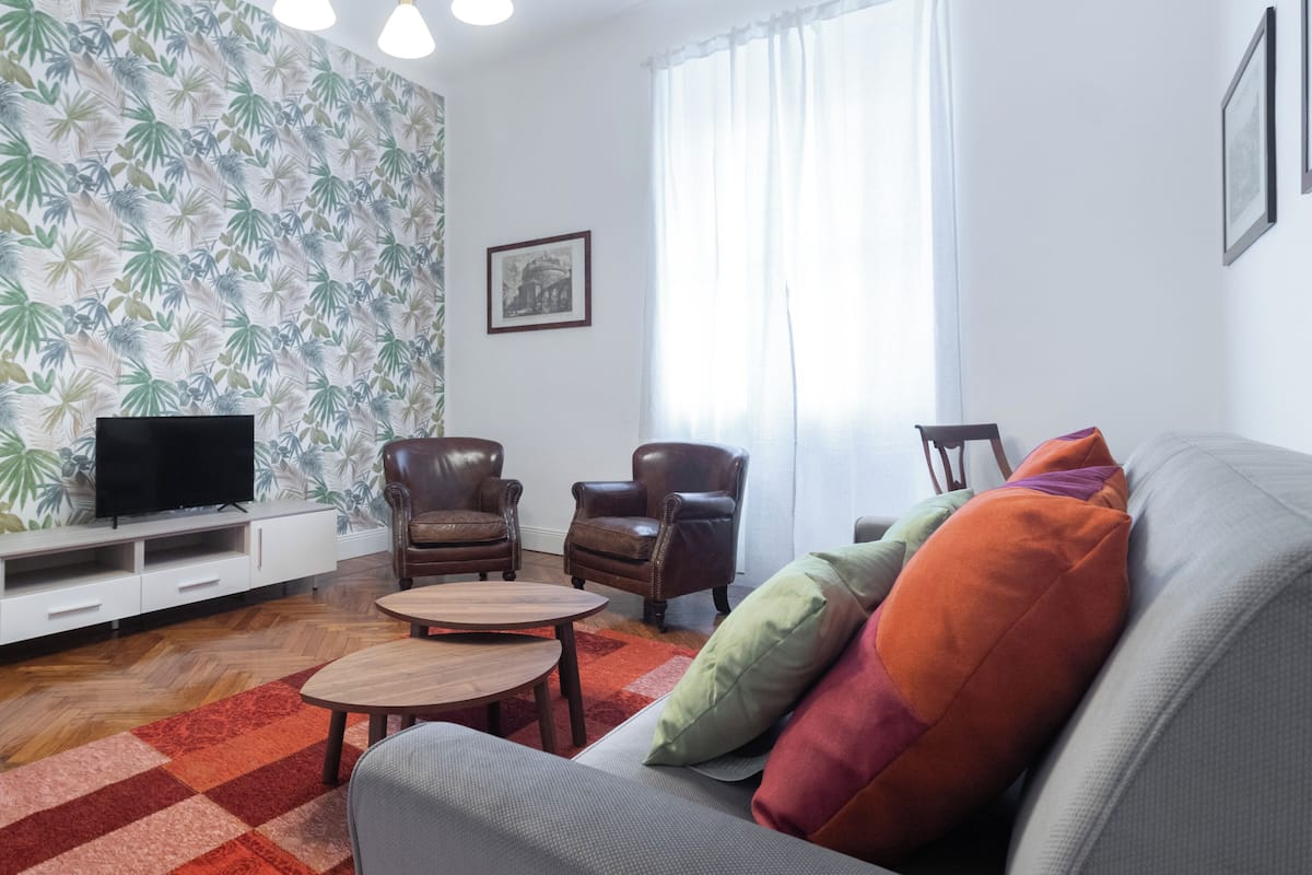 Un salón con papel tapiz estampado, un televisor sobre un mueble bajo, dos sillones de cuero, un sofá gris con cojines coloridos, una mesita redonda y una alfombra de cuadros rojos y naranjas.