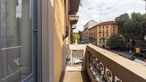 Un balcon doté d’une rambarde en métal, donnant sur une rue bordée de voitures garées et d’immeubles.