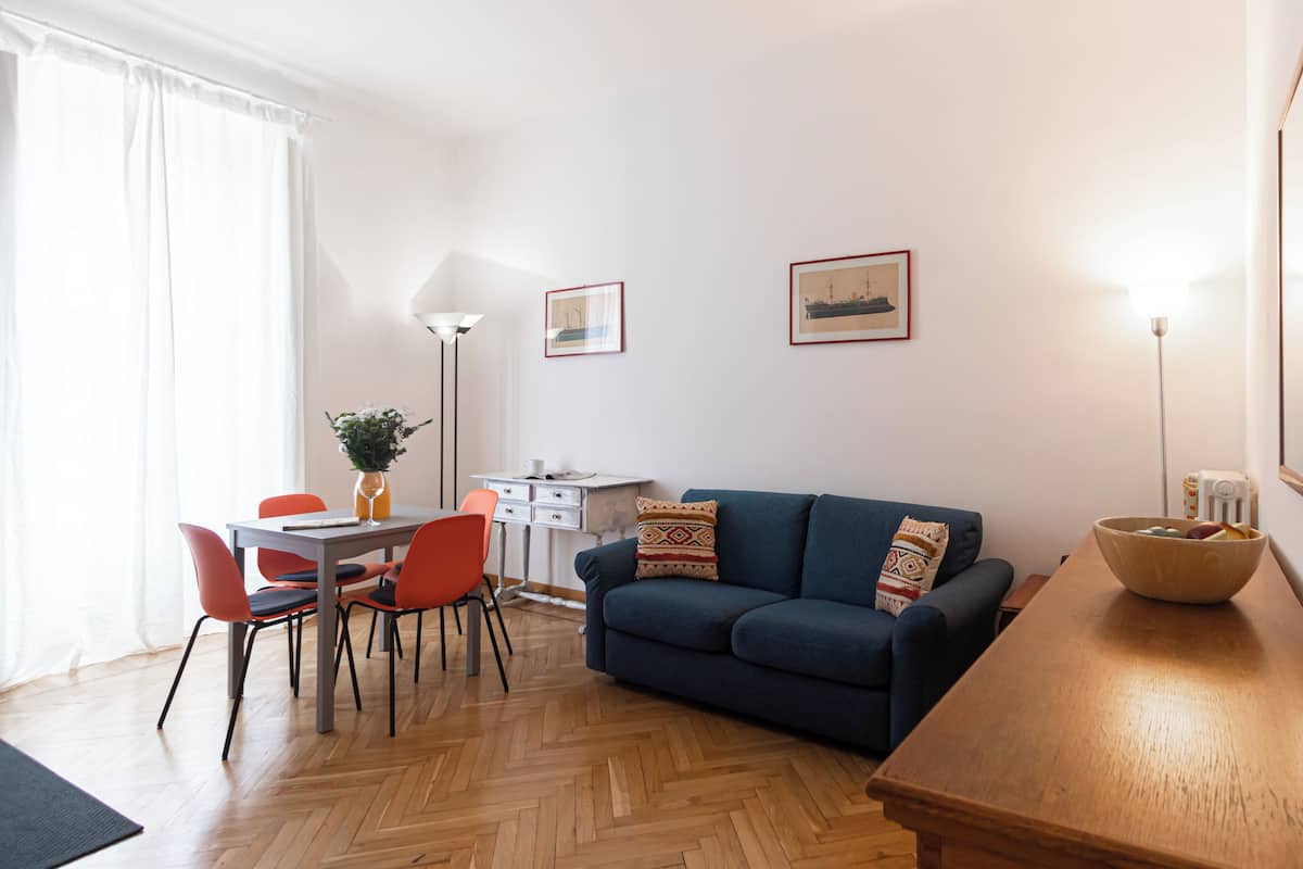 Una sala moderna con un sofá azul, una mesa de centro de madera y un comedor con una mesa blanca y sillas naranjas.