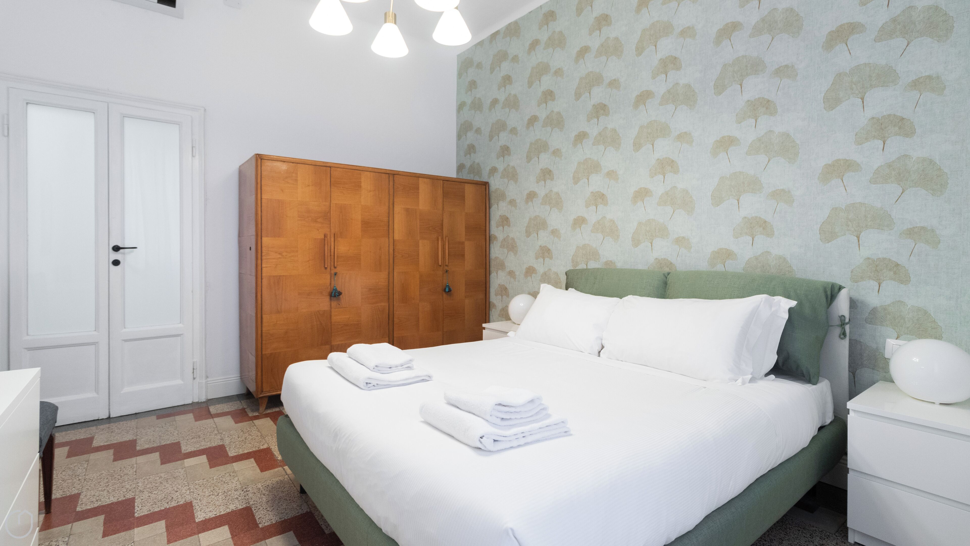 A neatly made bed with white linens and folded towels, a wooden wardrobe, a green headboard, and a patterned wallpaper.