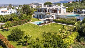 Eine große, weiße Villa mit Swimmingpool, umgeben von üppiger Vegetation und einer gebirgigen Kulisse.