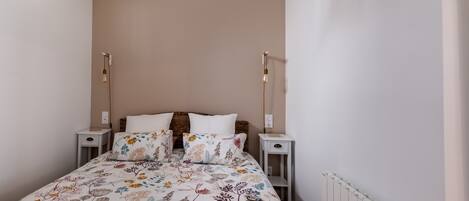 Ein Schlafzimmer mit blumigem Bettbezug, einer Lampenschirm aus Weidengeflecht, einem weißen Heizkörper und zwei Nachttischen mit Lampen.