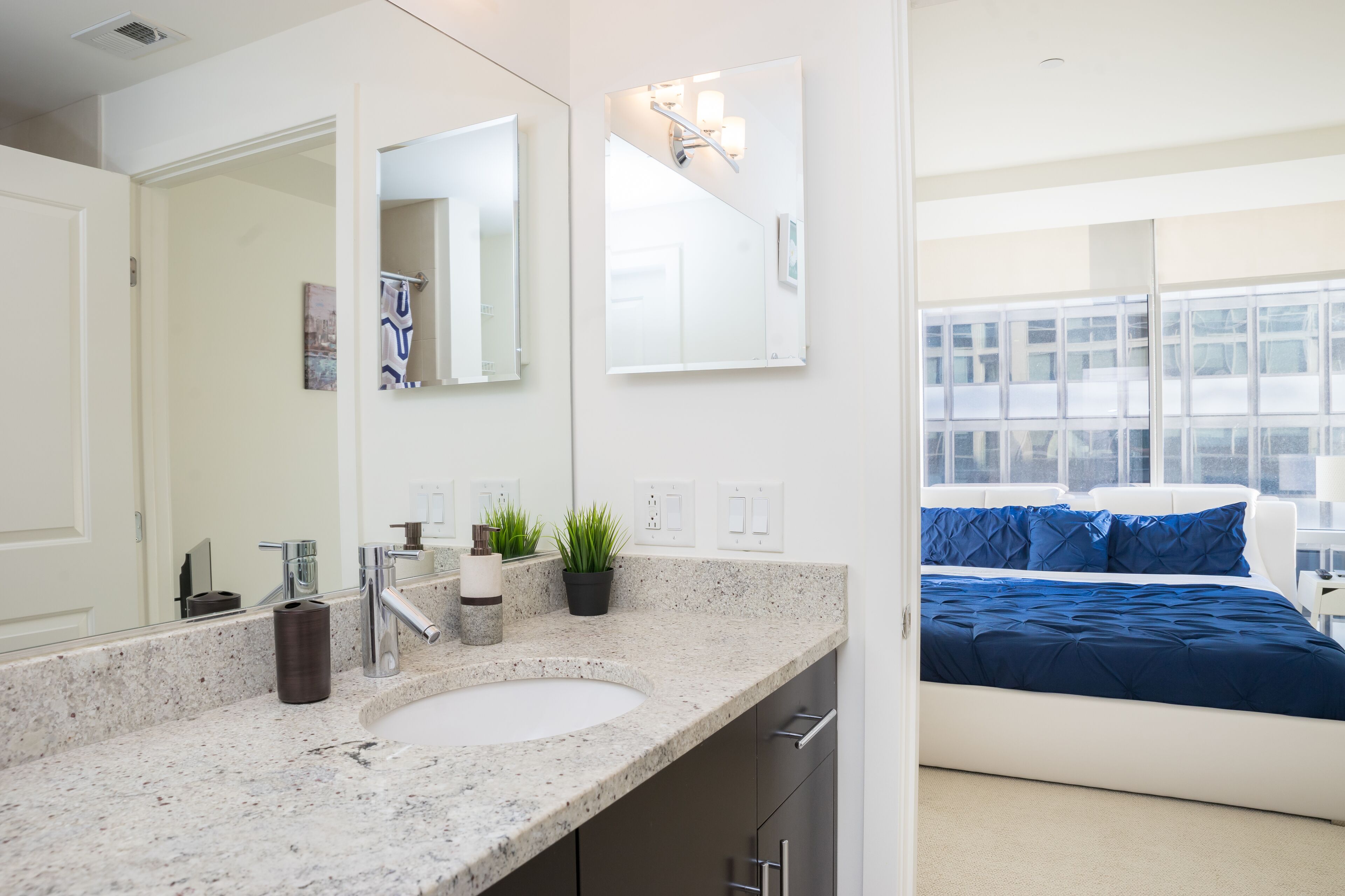 A modern bathroom with a granite countertop, a sink, and a mirror. There is a potted plant on the countertop and a bed with blue bedding in the adjacent room.